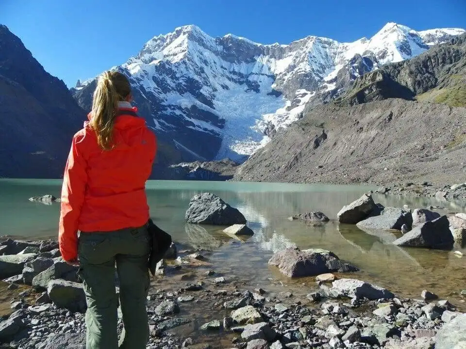 A woman is trekking in front of a lake with mountains in the background.
