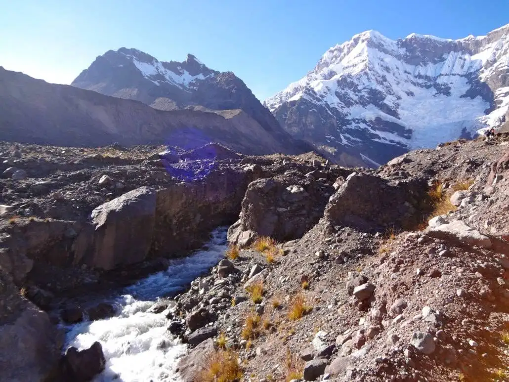 Apu Ausangate trekking spot showcases a stream meandering through a rocky area against the backdrop of majestic mountains.