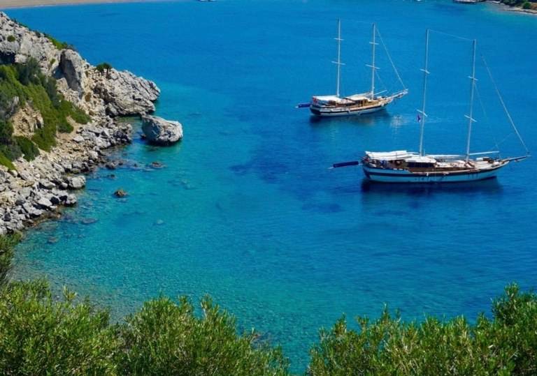 two sailboats are docked in the water near a rocky shore.