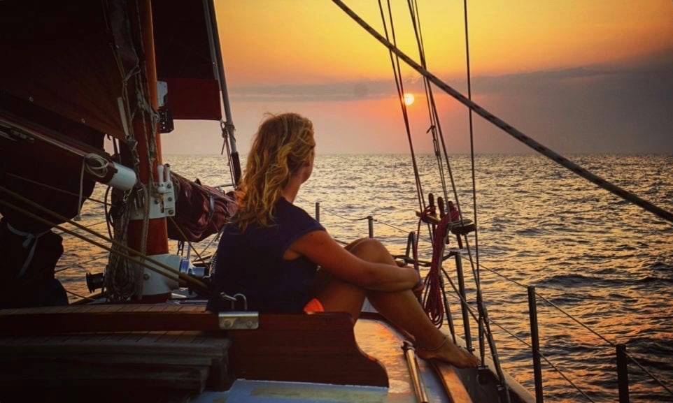 a woman sitting on the deck of a sailboat overlooking the ocean at sunset.
