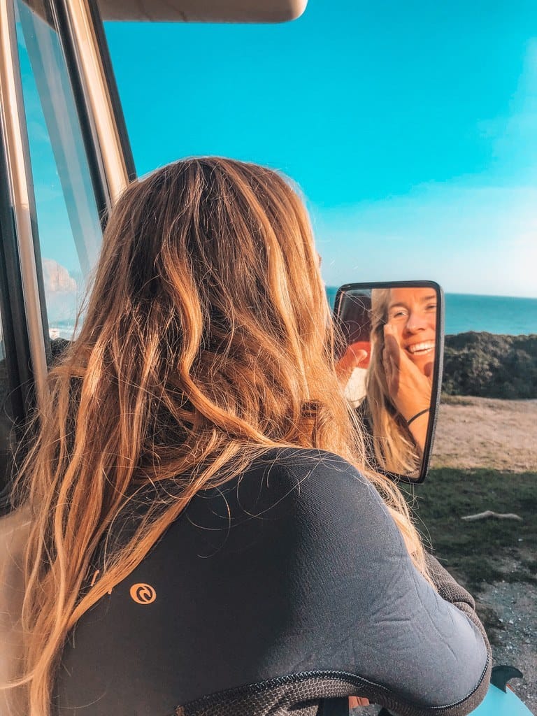 A woman with long blonde hair looks into a side mirror attached to a vehicle, smiling with a coastal landscape and blue sky in the background, her skin glowing from DIY mineral based sunscreen.
