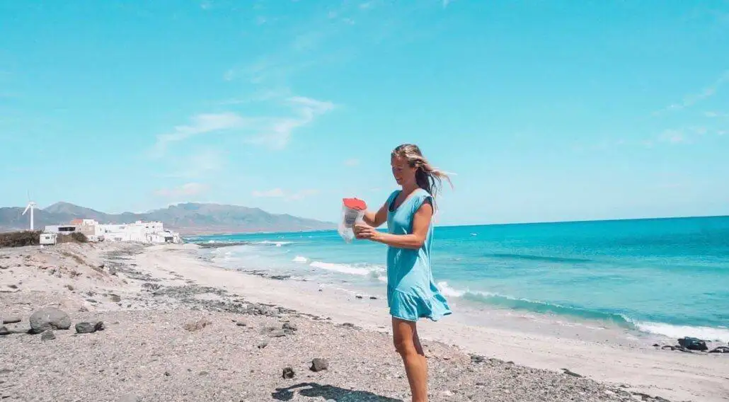 a woman in a blue dress standing on a beach, carrying a travel water filter.