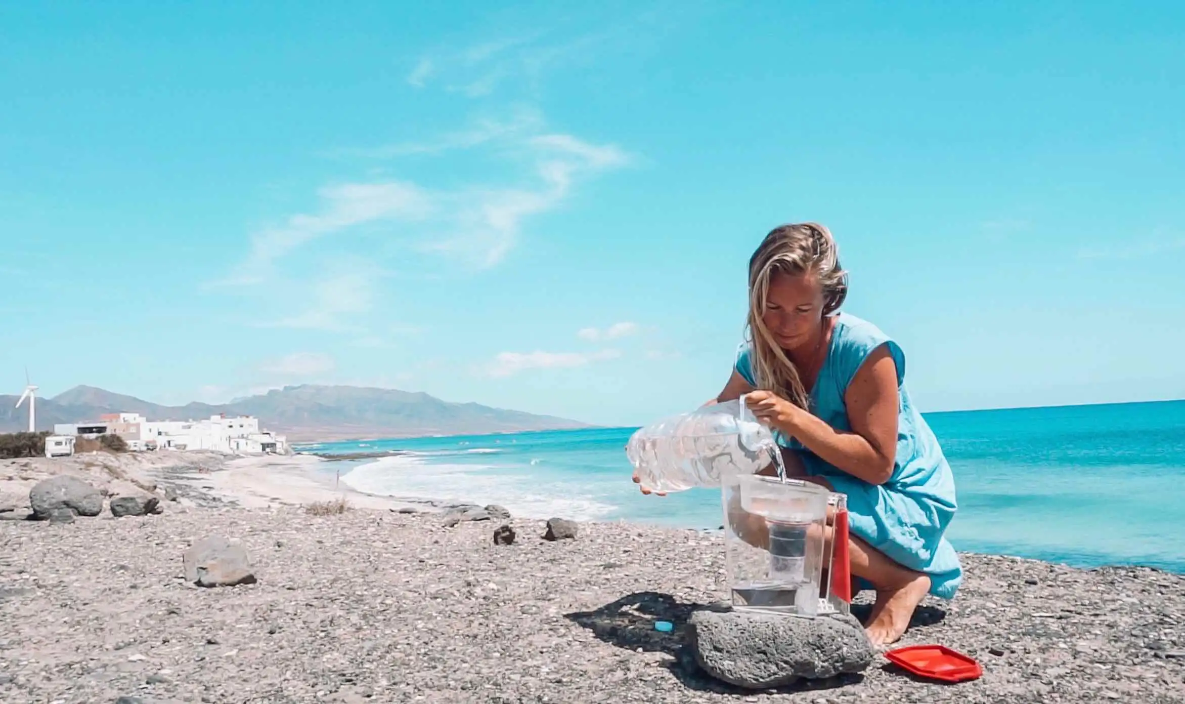 A woman utilizing a travel water filter while kneeling on the beach with a bucket of water.