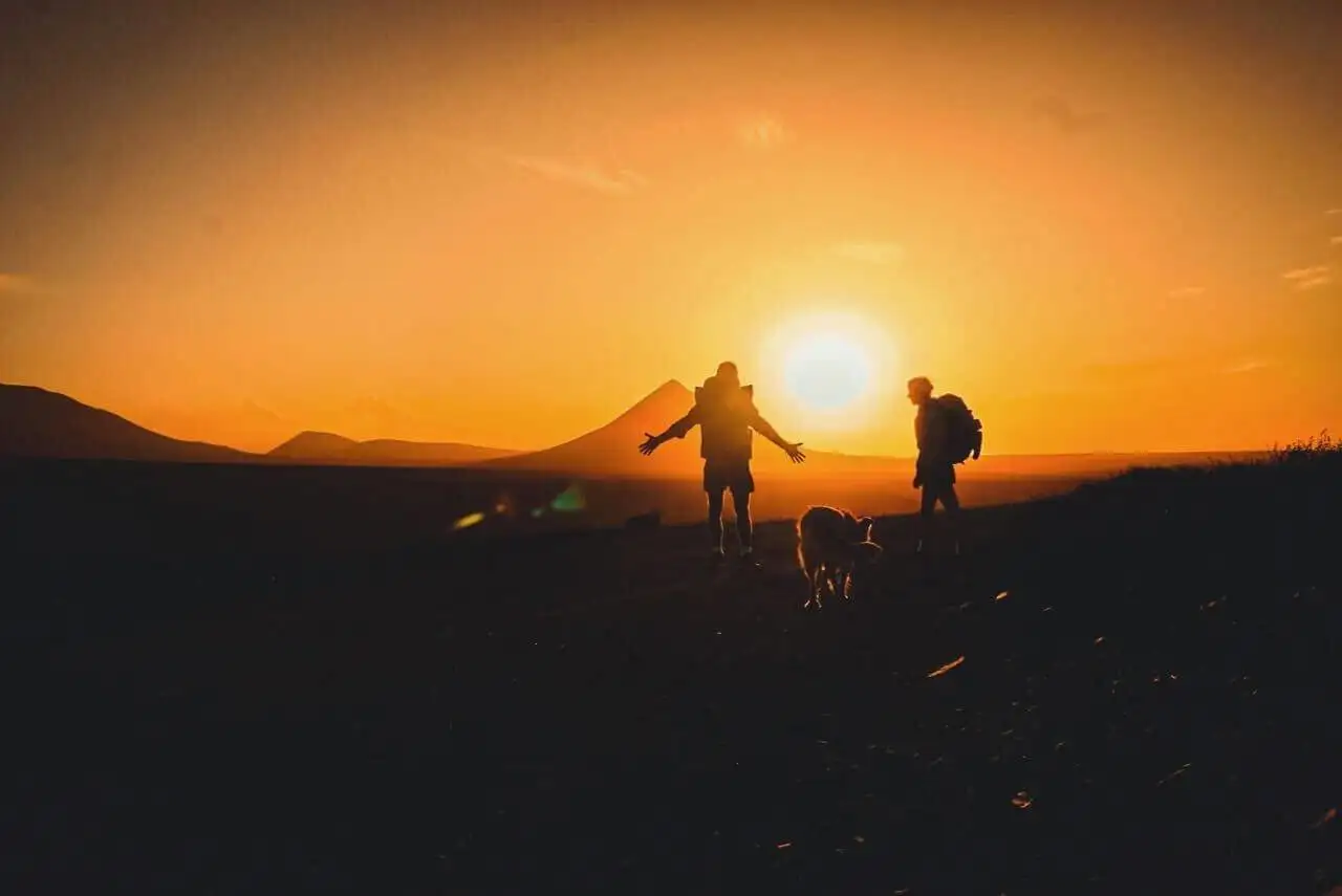 two people standing on a hill with a dog at sunset.
