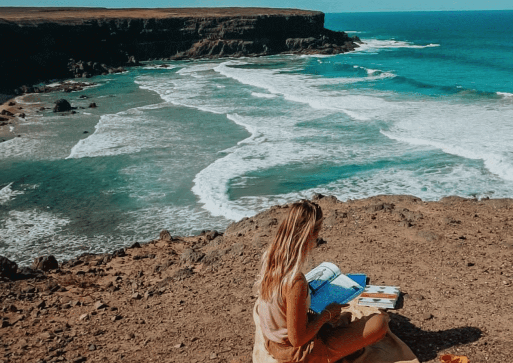 A woman reading a book on a cliff overlooking the ocean.