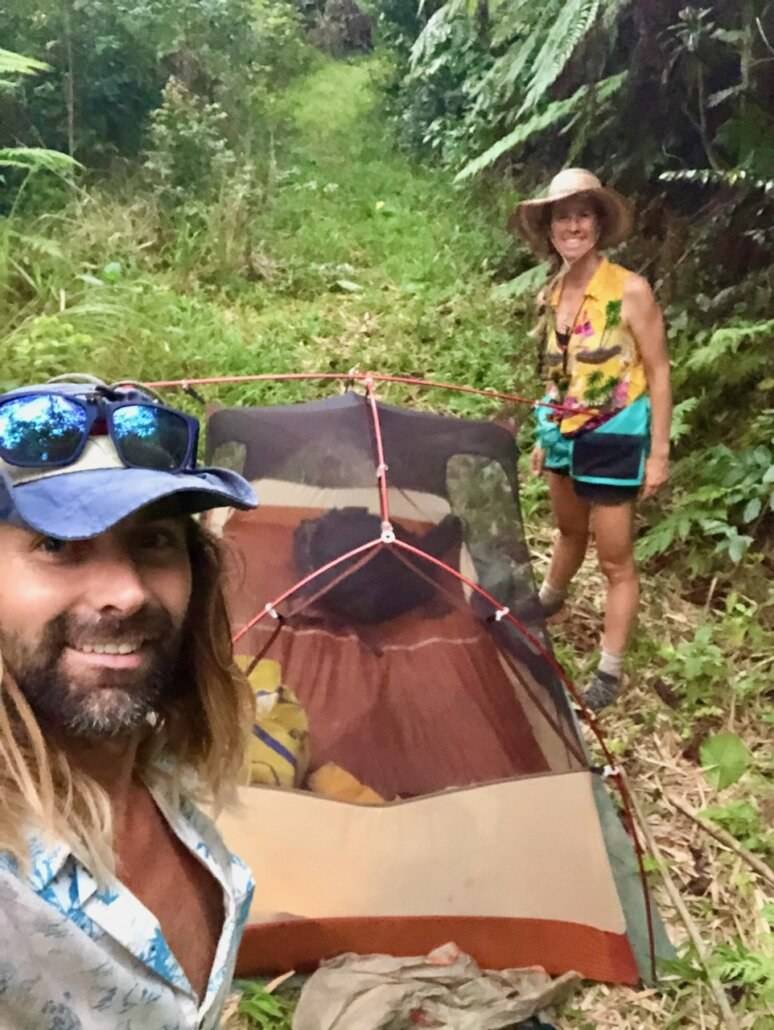 Two people standing next to a tent in the woods.