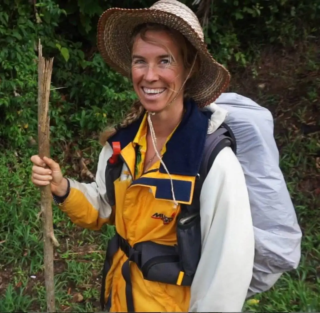 A woman wearing a hat and a backpack holding a stick.