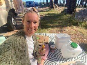 A woman smiling at the camera while sitting on a picnic blanket with camping gear and supplies around her in a wooded area.