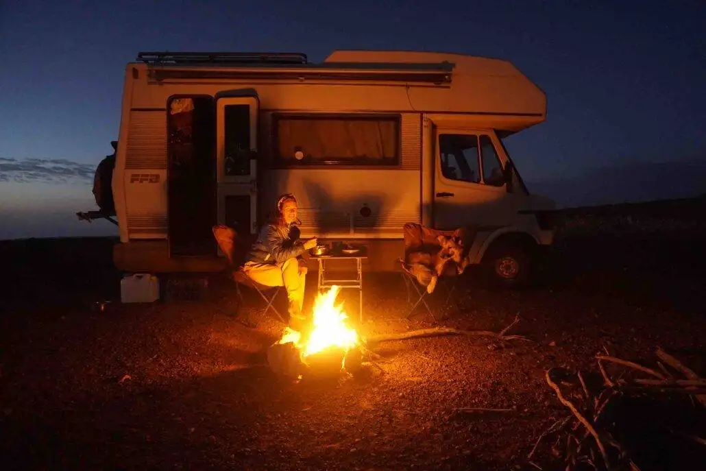 Person sitting by a campfire near a white camper van at dusk, seated on a folding chair with the Morocco & Western Sahara Vanlife Guide on the table next to them.