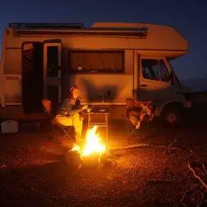 Person sitting by a campfire near a white camper van at dusk, seated on a folding chair with the Morocco & Western Sahara Vanlife Guide on the table next to them.