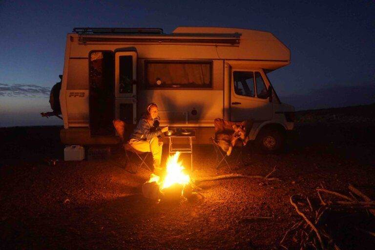 Person sitting by a campfire near a white camper van at dusk, seated on a folding chair with the Morocco & Western Sahara Vanlife Guide on the table next to them.