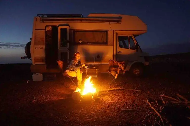 Person sitting by a campfire near a white camper van at dusk, seated on a folding chair with the Morocco & Western Sahara Vanlife Guide on the table next to them.