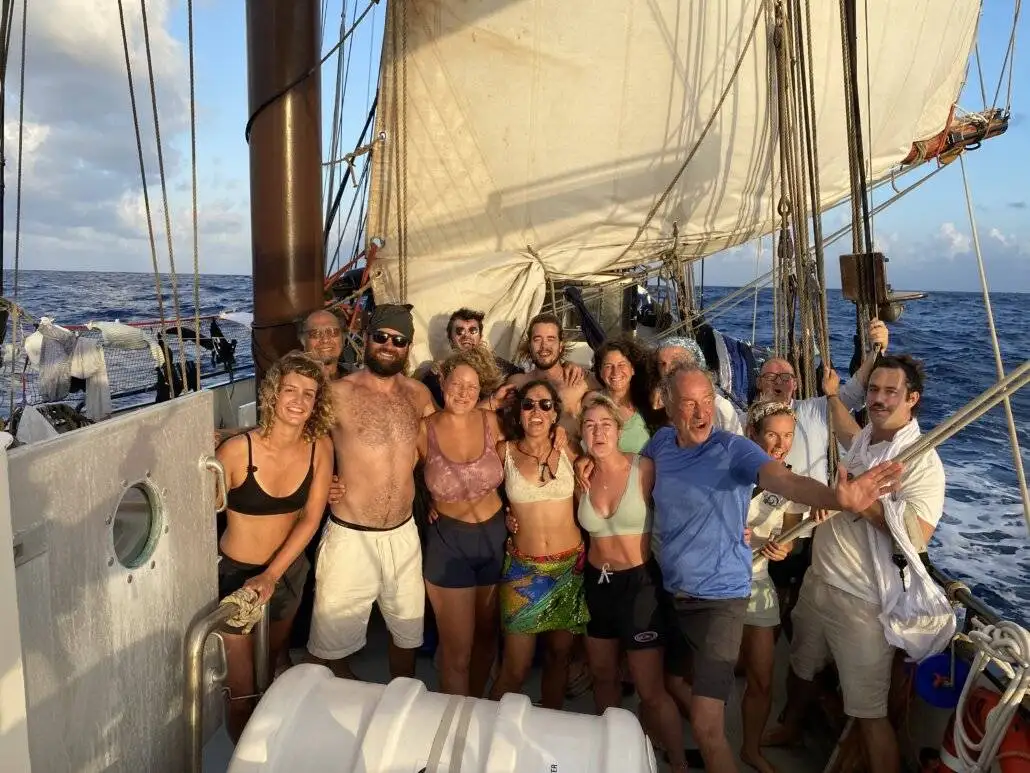 A group of people stand together smiling on the deck of a sailboat at sea, embracing outdoor adventure with sails raised and the ocean in the background.