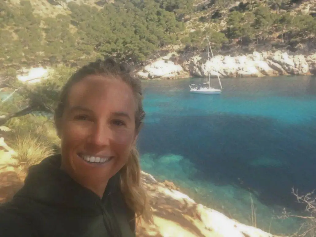 A person smiles at the camera on a rocky coastline with clear blue water and a sailboat anchored in the background, embracing nomadlife and adventure travel.