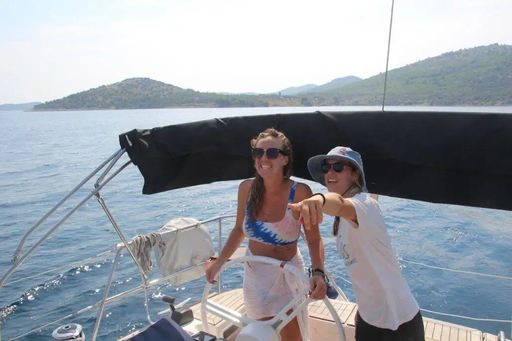 Two women on a sailboat enjoy outdoor adventure; one steers while the other points toward the water. Both wear sunglasses, framed by hilly coastline and calm sea—a perfect moment for those embracing nomadlife.