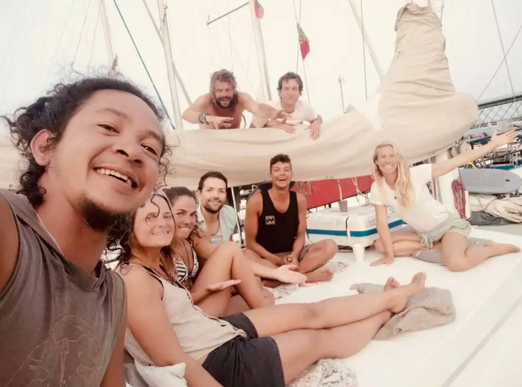 A group of seven people, embracing an alternative lifestyle, sit and smile on a sailboat, with sails and rigging visible in the background—capturing the spirit of adventure travel.