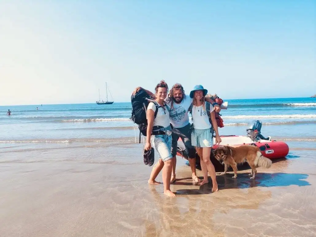 Three people and a dog embrace an outdoor adventure on a sunny beach near a red inflatable boat, with the sea and a sailboat in the background.