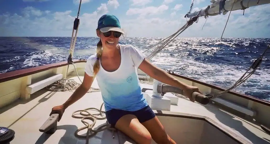 A woman wearing sunglasses and a cap sits at the helm of a sailboat on the open sea under a partly cloudy sky, embracing adventure travel and the freedom of an alternative lifestyle.