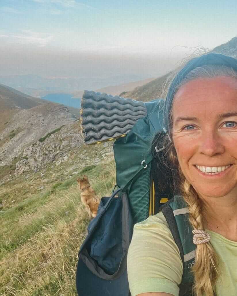 A woman with a backpack and braided hair smiles on a mountain trail; a dog sits on the grassy hillside behind her, overlooking a valley.