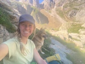 A natural nomad in a cap takes a selfie while sitting on rocky terrain with her dog beside her, mountains and sunlight in the background, capturing the spirit of outdoor adventure.