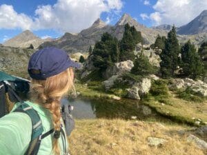 A woman with a braided ponytail and blue cap hikes near a scenic mountain range, standing by a small pond surrounded by rocks and trees.