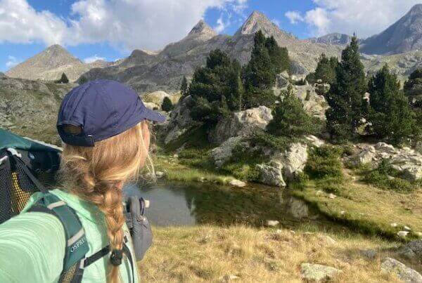 A woman with a braided ponytail and blue cap hikes near a scenic mountain range, standing by a small pond surrounded by rocks and trees.