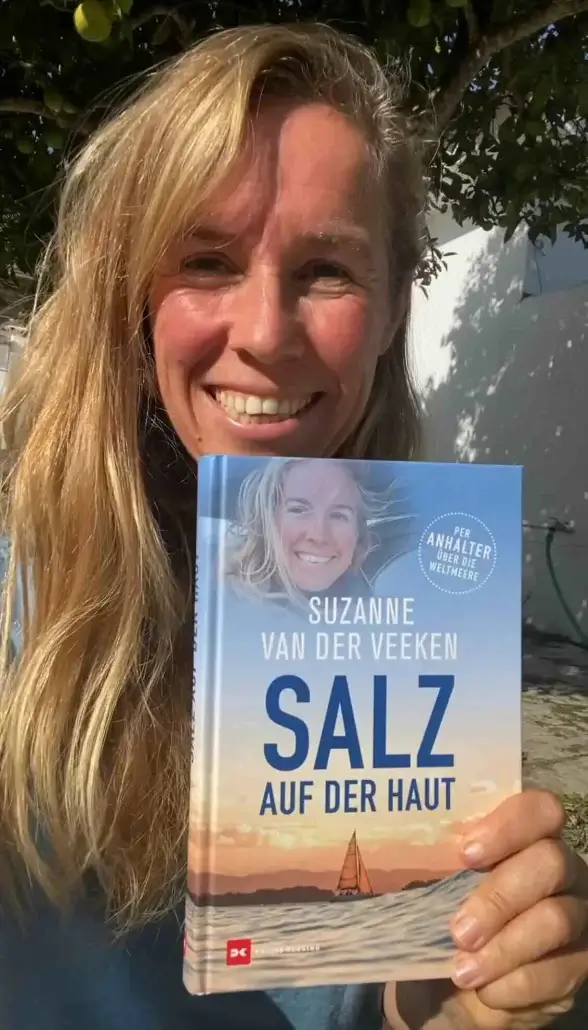 A person smiling while holding up the book "Salz auf der Haut" by Suzanne van der Veeken outdoors.