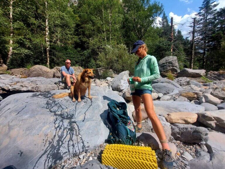 Two people and a dog rest on large rocks in a forested area. One person is seated, and the other stands with a water bottle. A backpack and sleeping mat are on the ground.