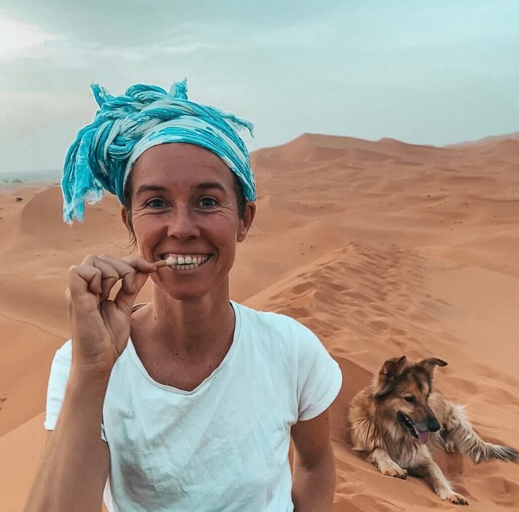 Person in a white shirt and blue headscarf stands in a desert holding a twig to their teeth, with a dog lying down in the sand nearby.