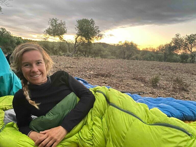 Person lying in a bright green sleeping bag outdoors, surrounded by trees, with a cloudy sky and the sun setting in the background.