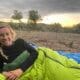 Person lying in a bright green sleeping bag outdoors, surrounded by trees, with a cloudy sky and the sun setting in the background.