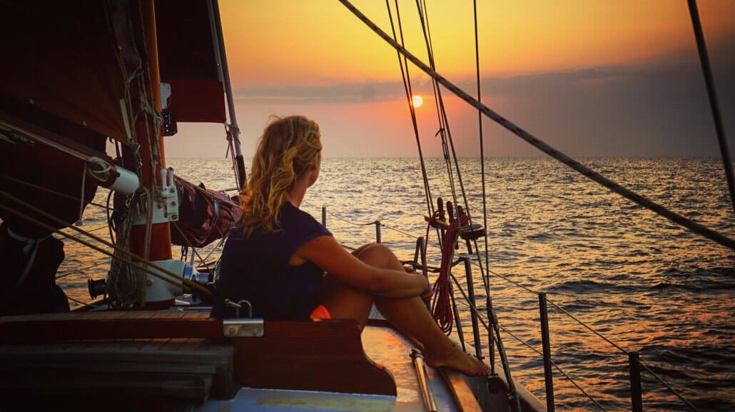 A person sits on a sailboat, looking at the sunset over the ocean, with orange and pink hues in the sky.