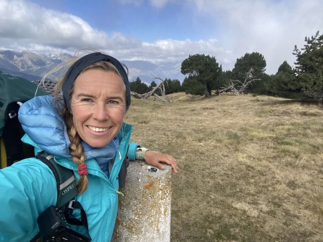 A person in outdoor clothing takes a selfie on a grassy mountain landscape with trees and cloudy sky in the background.