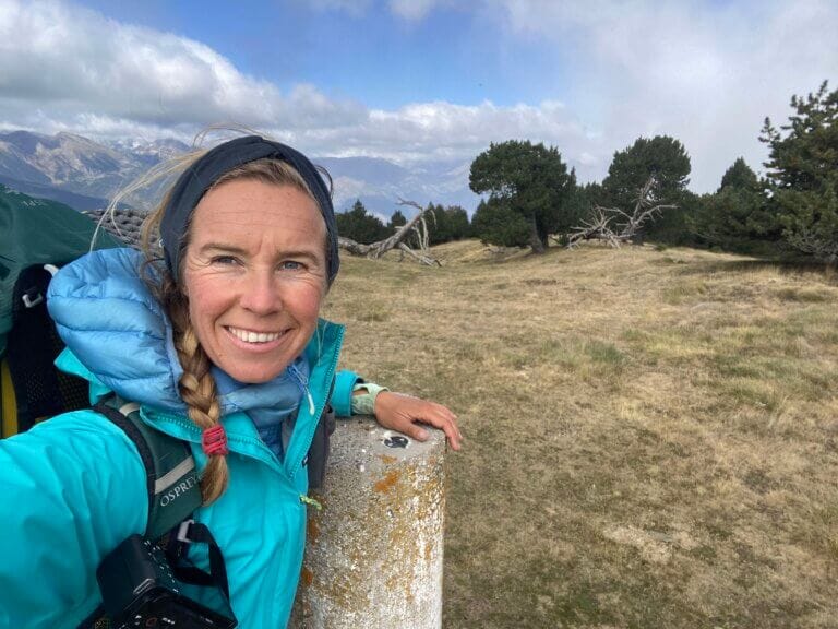 A person in outdoor clothing takes a selfie on a grassy mountain landscape with trees and cloudy sky in the background.
