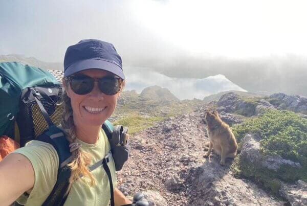 A woman wearing a cap, sunglasses, and backpack takes a selfie while hiking on a rocky hilltop; a dog sits nearby, with misty mountains in the background.