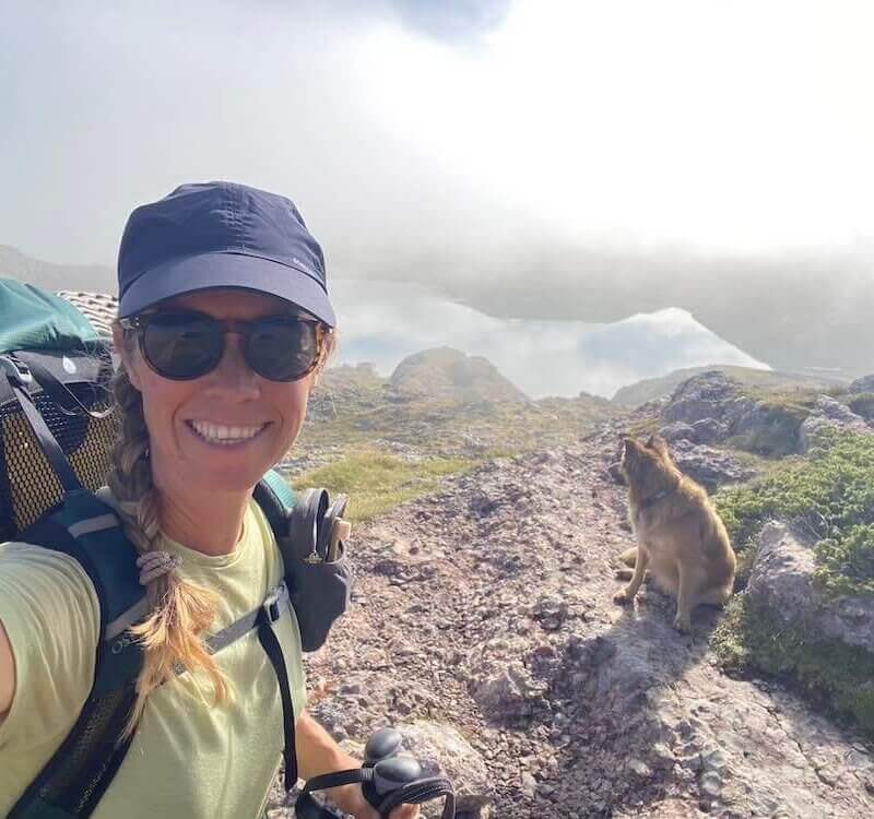 A woman wearing a cap, sunglasses, and backpack takes a selfie while hiking on a rocky hilltop; a dog sits nearby, with misty mountains in the background.