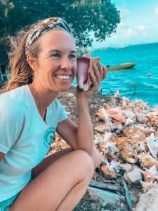 A woman kneels by a pile of conch shells near the water, enjoying an outdoor adventure as she holds one shell to her ear and smiles, with trees and a boat in the background.