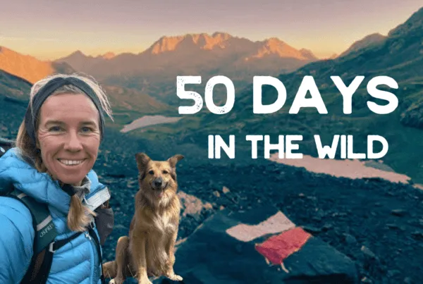 A woman in hiking gear and a dog pose on a rocky mountain trail at sunrise with "50 Days in the Wild" text overlaid on the image.