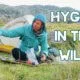A person in outdoor gear sits by a tent in a grassy area, pouring water from a bottle. Large text reads, "Hygiene in the Wild.