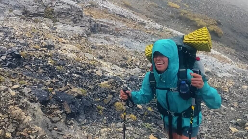 A hiker wearing a teal rain jacket and large backpack with yellow sleeping pad ascends a rocky, mountainous trail in wet conditions.