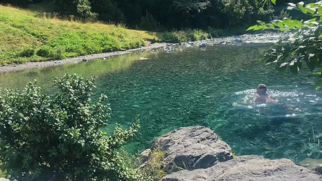 Clear river water with visible rocks, green foliage on the banks, and a person swimming near the right edge of the image.