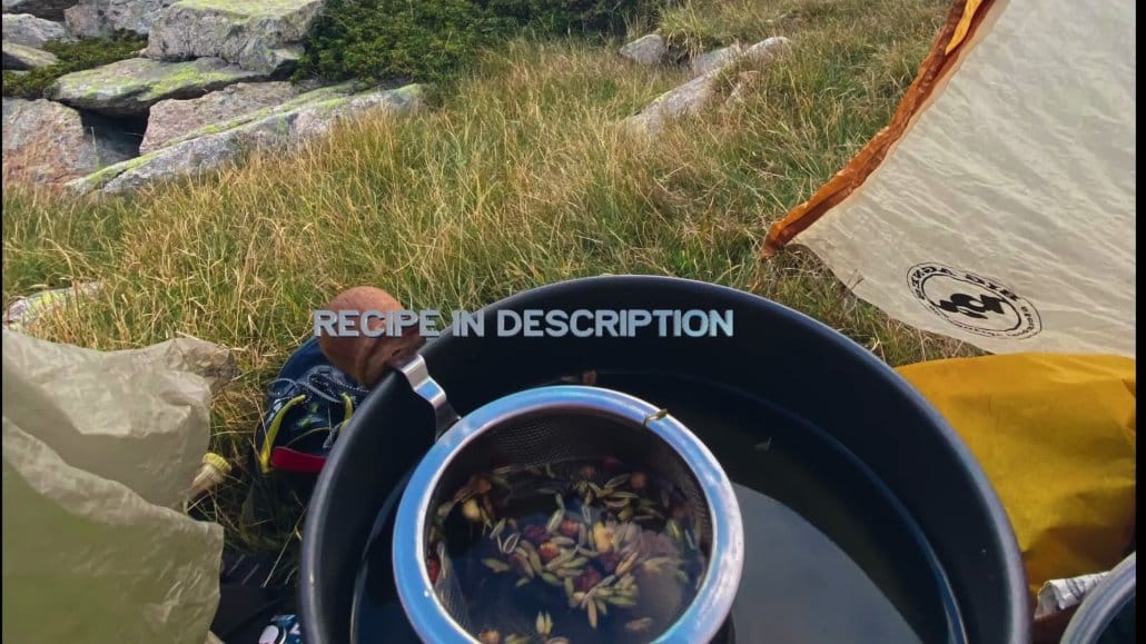 A metal strainer with herbs sits in a pot of water on grass near camping gear and a tent; outdoor rocks and greenery are visible in the background.