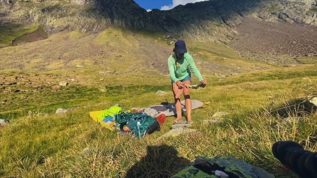 A person in outdoor clothing stands on grassy terrain with camping gear, surrounded by mountains under a partly cloudy sky.