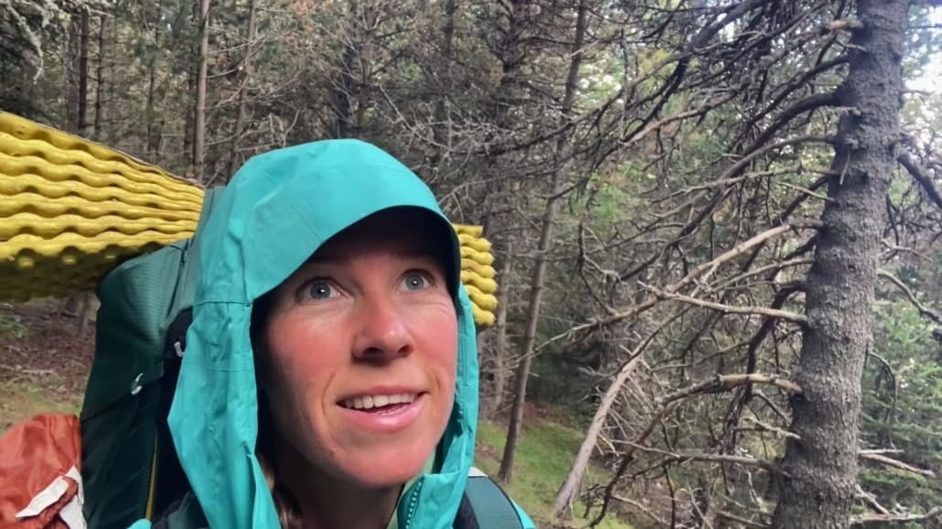 A person wearing a teal hooded jacket and backpack stands in a forest, looking upward; yellow sleeping pad visible on the backpack.