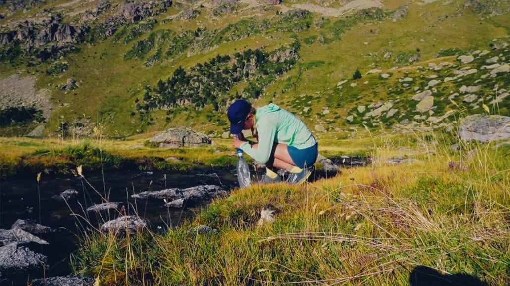 Person crouching by a stream in a grassy mountainous area, collecting water with a container on a sunny day.