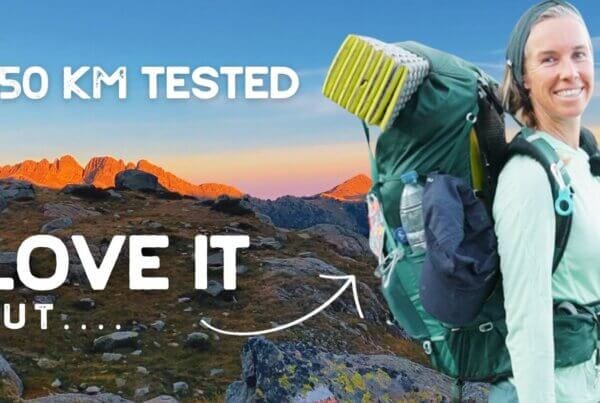 A woman with a large hiking backpack stands on a rocky mountain trail at sunset. Text reads: "850 KM TESTED. LOVE IT BUT....
