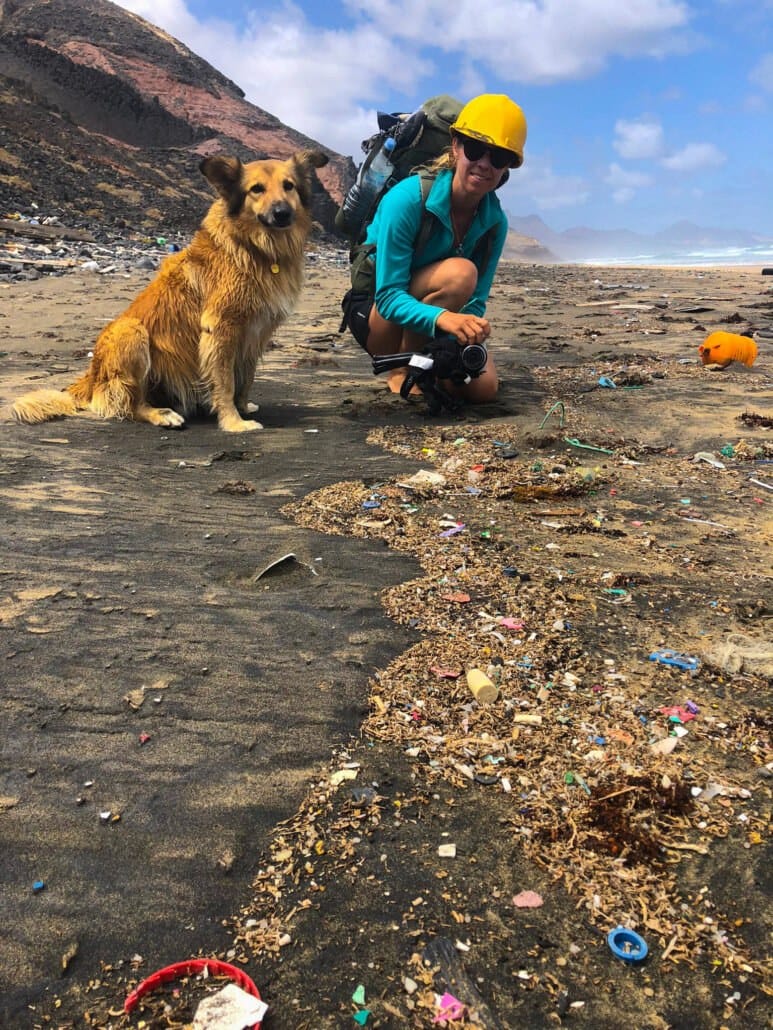 A woman wearing a yellow hard hat and backpack kneels on a littered beach with a dog beside her; scattered plastic debris covers the sand.