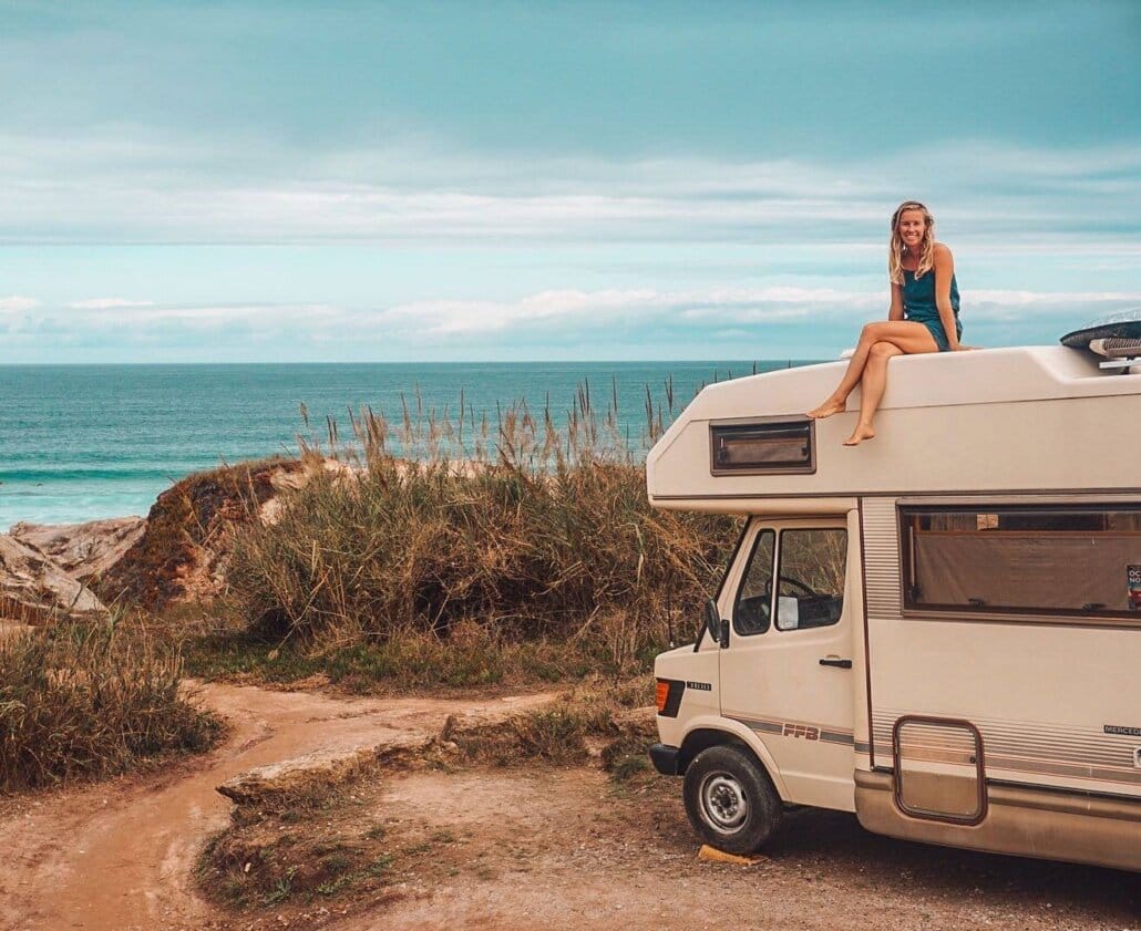A person sits on top of a camper van parked near the coast, with grass, dirt path, and the ocean visible in the background.