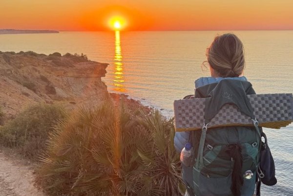 A person with a backpack and a small dog stand on a coastal trail at sunset, overlooking the ocean with the sun low on the horizon.
