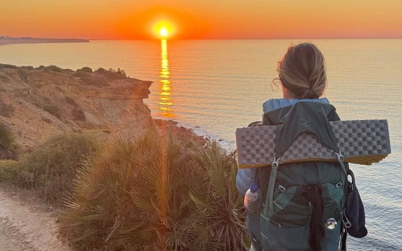 A person with a backpack and a small dog stand on a coastal trail at sunset, overlooking the ocean with the sun low on the horizon.